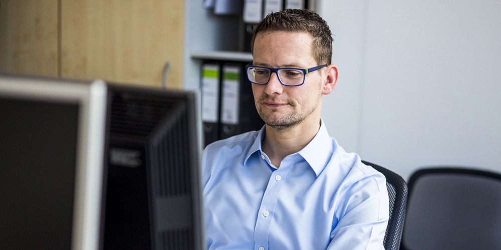 Mann mit Brille und blauem Hemd sitzt an seinem Arbeitsplatz und schaut konzentiert auf den Computermonitor.