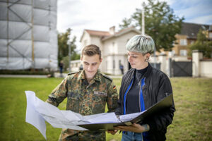 Christina schaut zusammen mit einem Soldaten der Bundeswehr auf Baupläne. Sie stehen auf einer Wiese vor einem eingerüsteten Gebäude, welches von der Bundeswehr genutzt wird und dessen Sanierung Christina betreut.