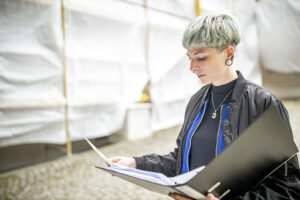 Christina mit einem Aktenordner in der Hand. Sie schaut konzentriert auf die Papiere. Im Hintergrund ist ein Baugerüst zu sehen.