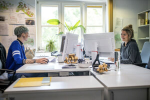 Christina im Büro an ihrem Arbeitsplatz mit Computer. Ihr gegenüber sitzt ihre Kollegin.