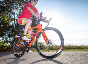 Rennradfahrerin mit Helm und Sonnenbrille auf einem orangenen Rennrad fährt seitlich nach rechts aus dem Bild.