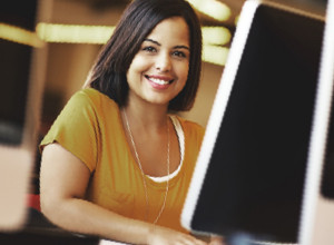 Lächelnde junge Frau mit senfgelbem T-Shirt schaut seitlich hinter einem Computermonitor hervor.