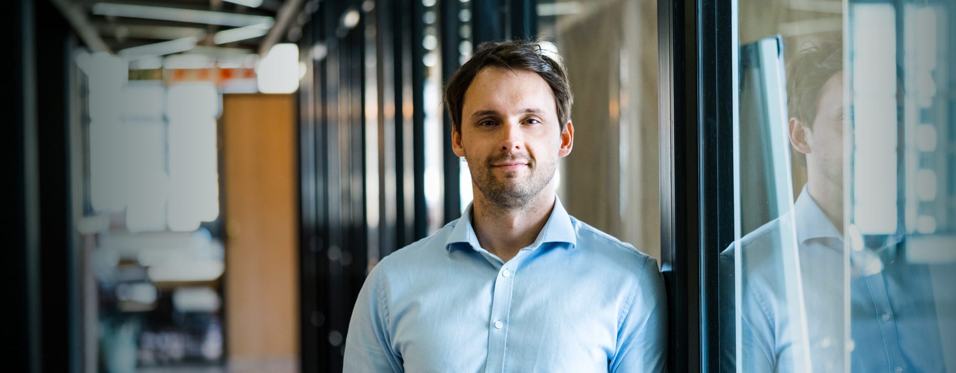 Verwaltungsmitarbeiter Mann mit blauem Hemd lehnt lässig an einer modernen, großen Fensterfront und schaut selbstbewusst in die Kamera.