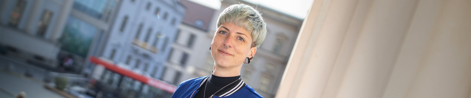Projektleiterin Bau Blonde, junge Frau mit Kurzhaarschnitt, Nasenring und Flesh Tunneln in den Ohrläppchen schaut freundlich in die Kamera. Sie heißt Christina und arbeitet beim Brandenburgischen Landesbetrieb für Liegenschaften und Bauen als Projektleiterin.